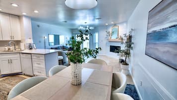 A modern kitchen with white cabinets, a dining area with a wooden table and chairs, and a living room with a fireplace and a large painting.