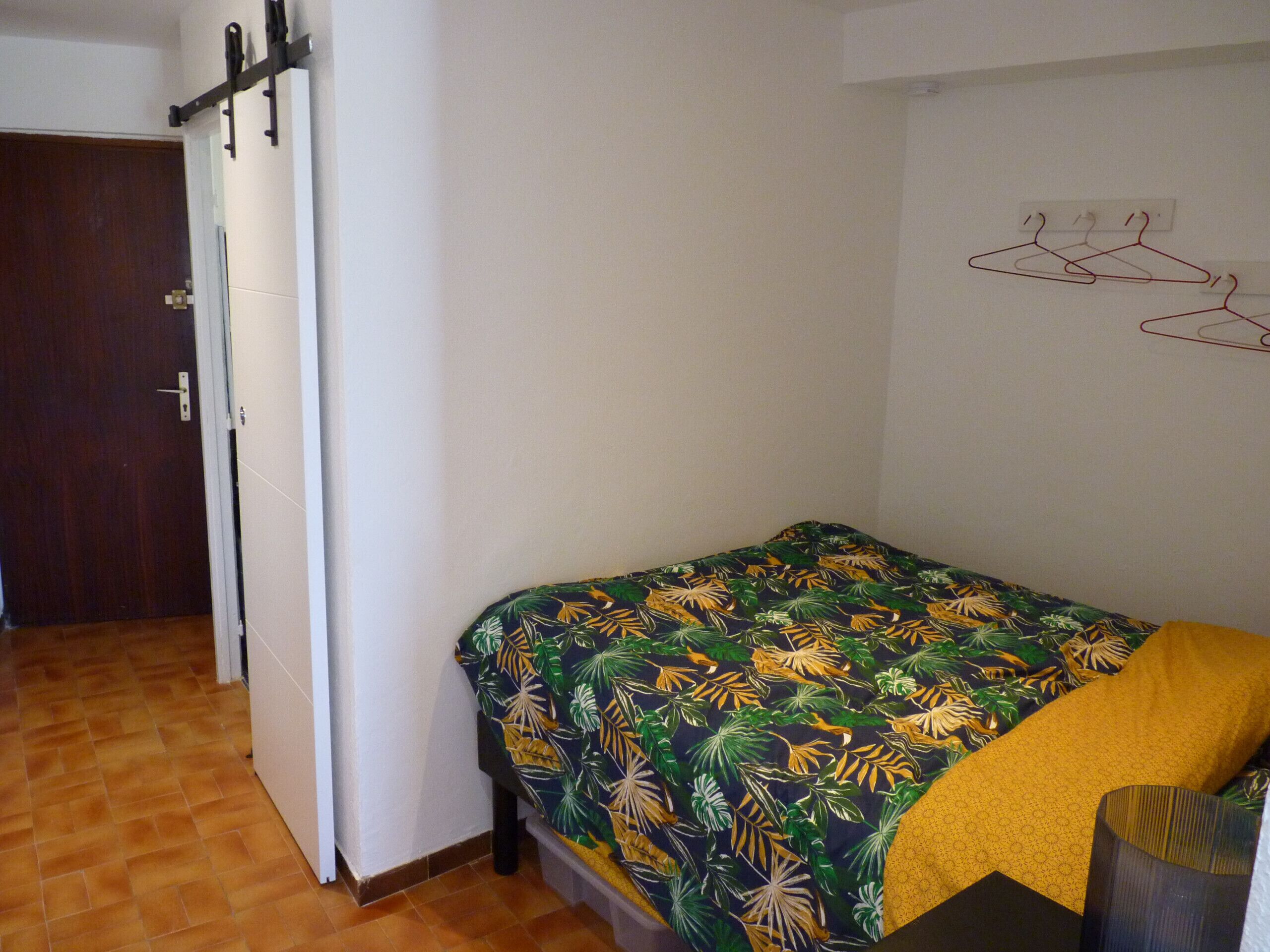 A small, single-bed room with a tropical-themed bedspread, a wooden door, and a wall-mounted coat rack.
