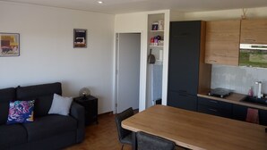 Una sala de estar moderna con un sofá oscuro, una mesa de comedor de madera y una zona de cocina con armarios de madera y un horno empotrado.