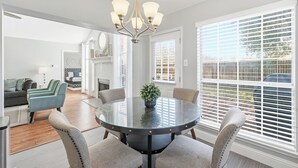 Ein Essbereich mit einem runden Tisch, Stühlen und einer Chandelier. Ein Kamin und ein Blick ins Freie durch große Fenster sind vorhanden.