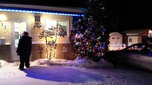 Een huis met een sneeuwbedekte voortuin, een kerstboom versierd met kleurrijke lichtjes, en een auto op de oprit.