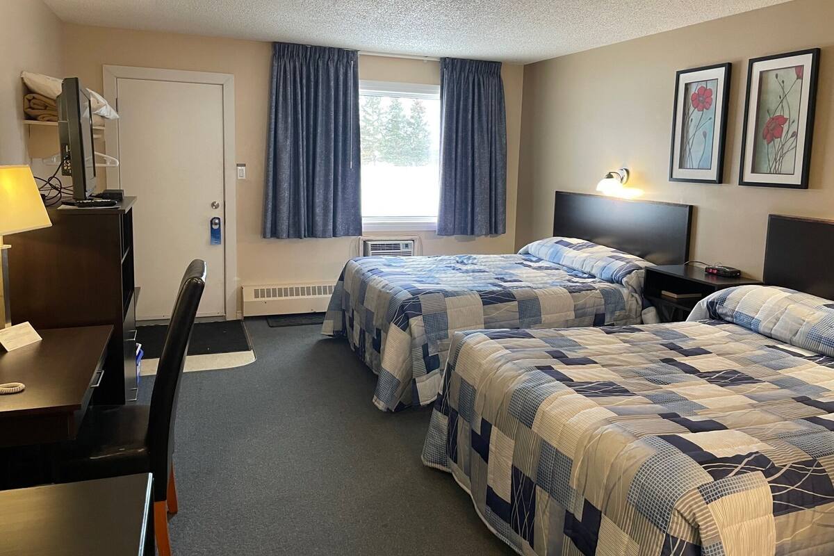 Quarto de hotel com duas camas, uma mesa, uma cadeira, janela com cortinas e um radiador.