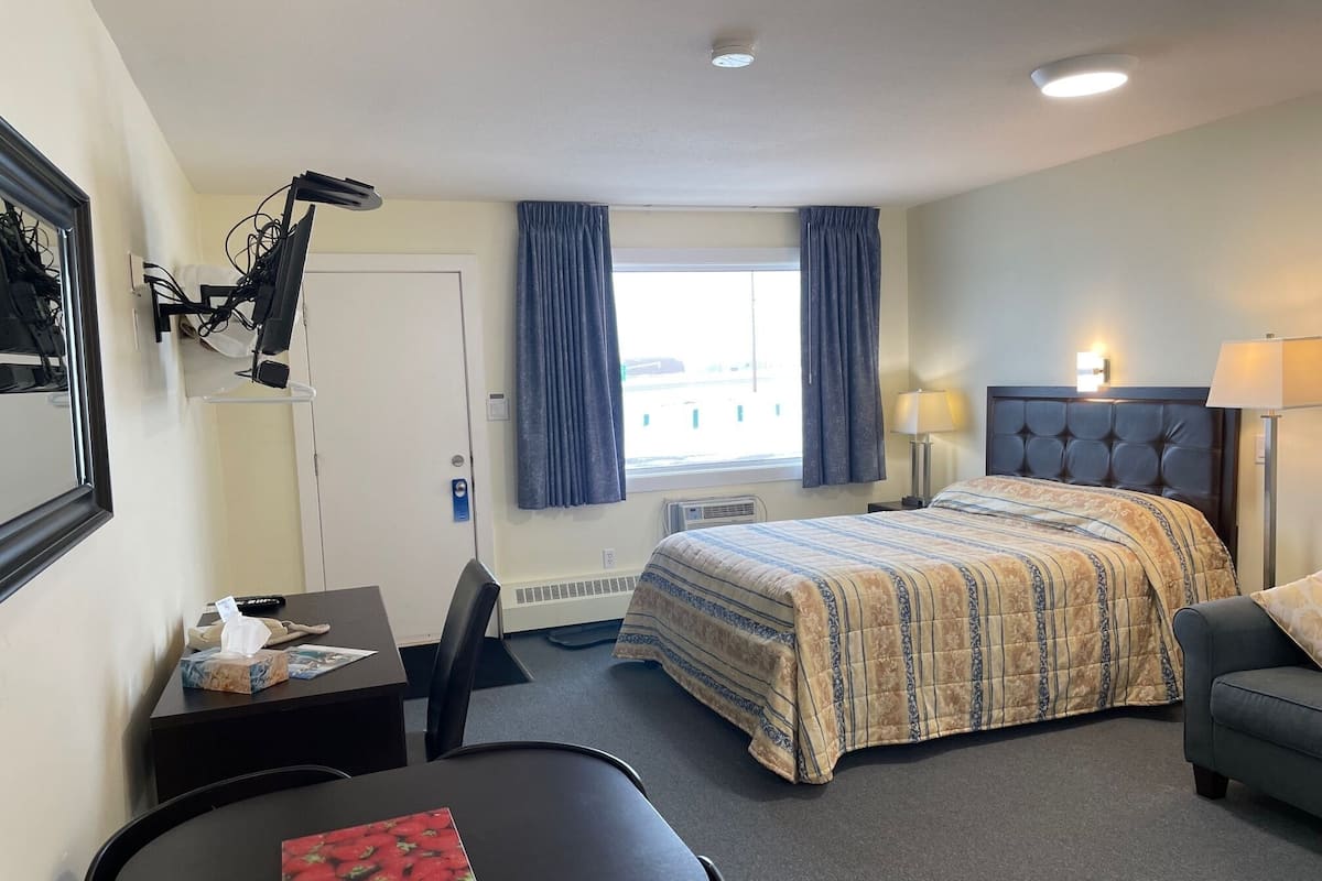Quarto de hotel com cama, escrivaninha, cadeira, luminária e janela com cortinas.