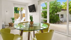 A modern dining area with a glass table and yellow chairs, a potted plant, and a view of a garden through large sliding doors. - Boca Catalina Beach 1 Bed/Kitchen Private Pool (Noord)