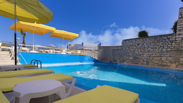 Un área de piscina con sillones de descanso amarillos y blancos, un muro de piedra y un cielo azul despejado.