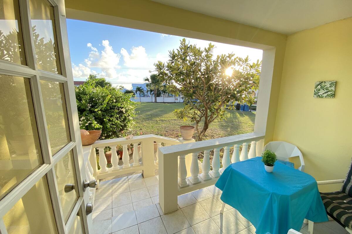 A balcony with a view of a garden, a table with a blue tablecloth, and a chair.