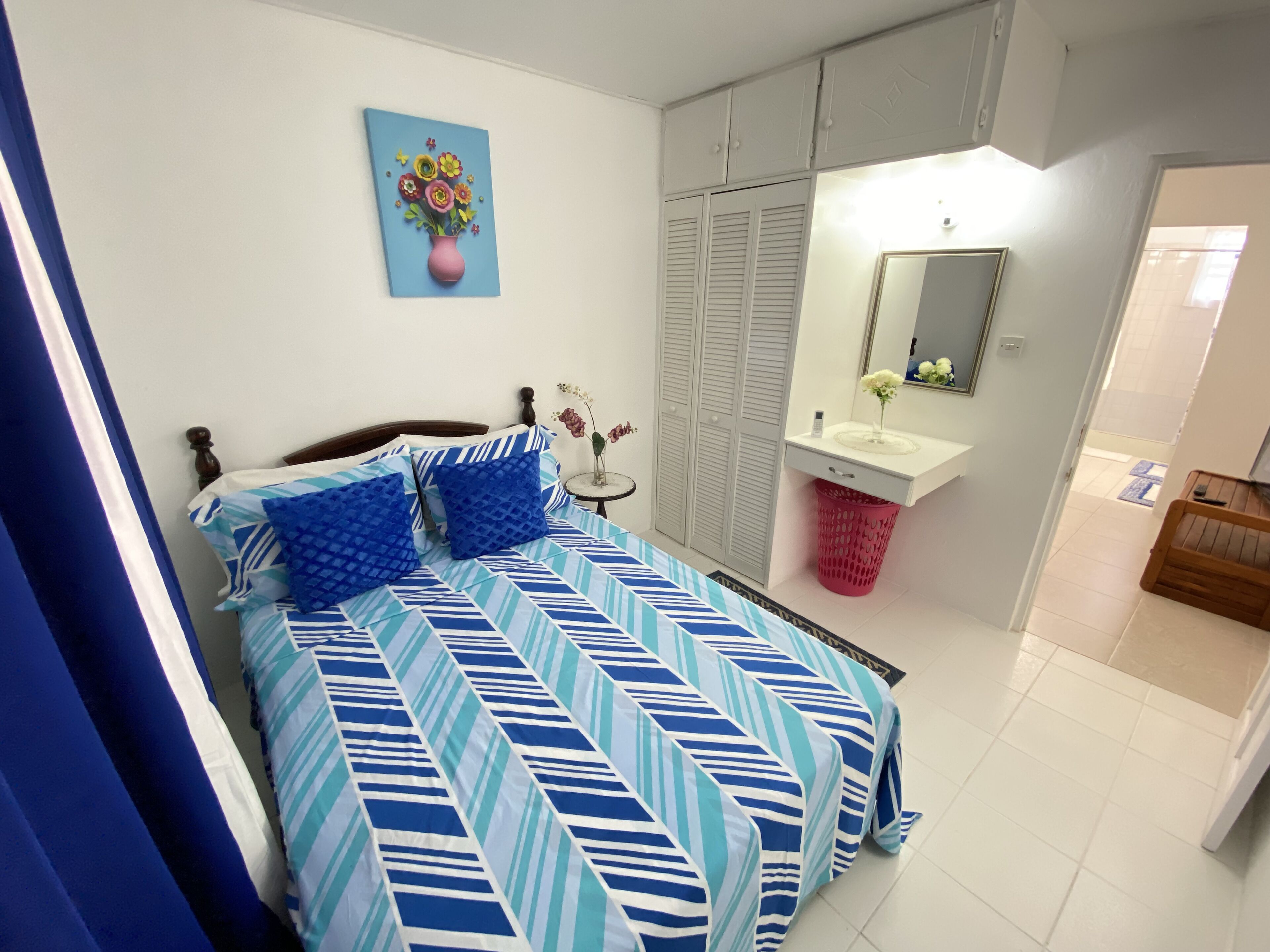 A bedroom with a bed, a bedside table with a vase, a wardrobe, a bathroom with a sink and mirror, and a door leading to another room.