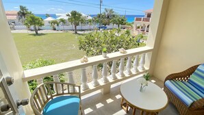 A balcony with a wicker chair, a small round table, and a view of the ocean and buildings. - Searocks Beach Apartment (Oistins)
