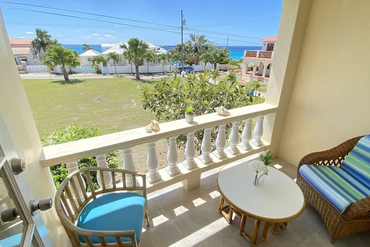 A balcony with a wicker chair, a small round table, and a view of the ocean and buildings.