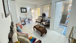 A living room with a sofa, chairs, a coffee table, a dining table with chairs, a fan, and a staircase leading to an upper floor. - Searocks Beach Apartment (Oistins)