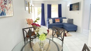 A modern living room with a blue sofa, a glass dining table with chairs, and a ceiling fan. - Searocks Beach Apartment (Oistins)