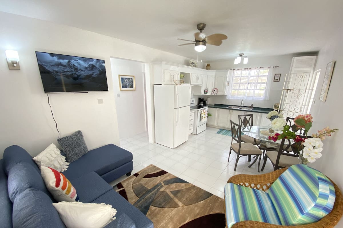 A modern living room with a sofa, a dining table with chairs, and a kitchen area with white cabinets and a refrigerator.