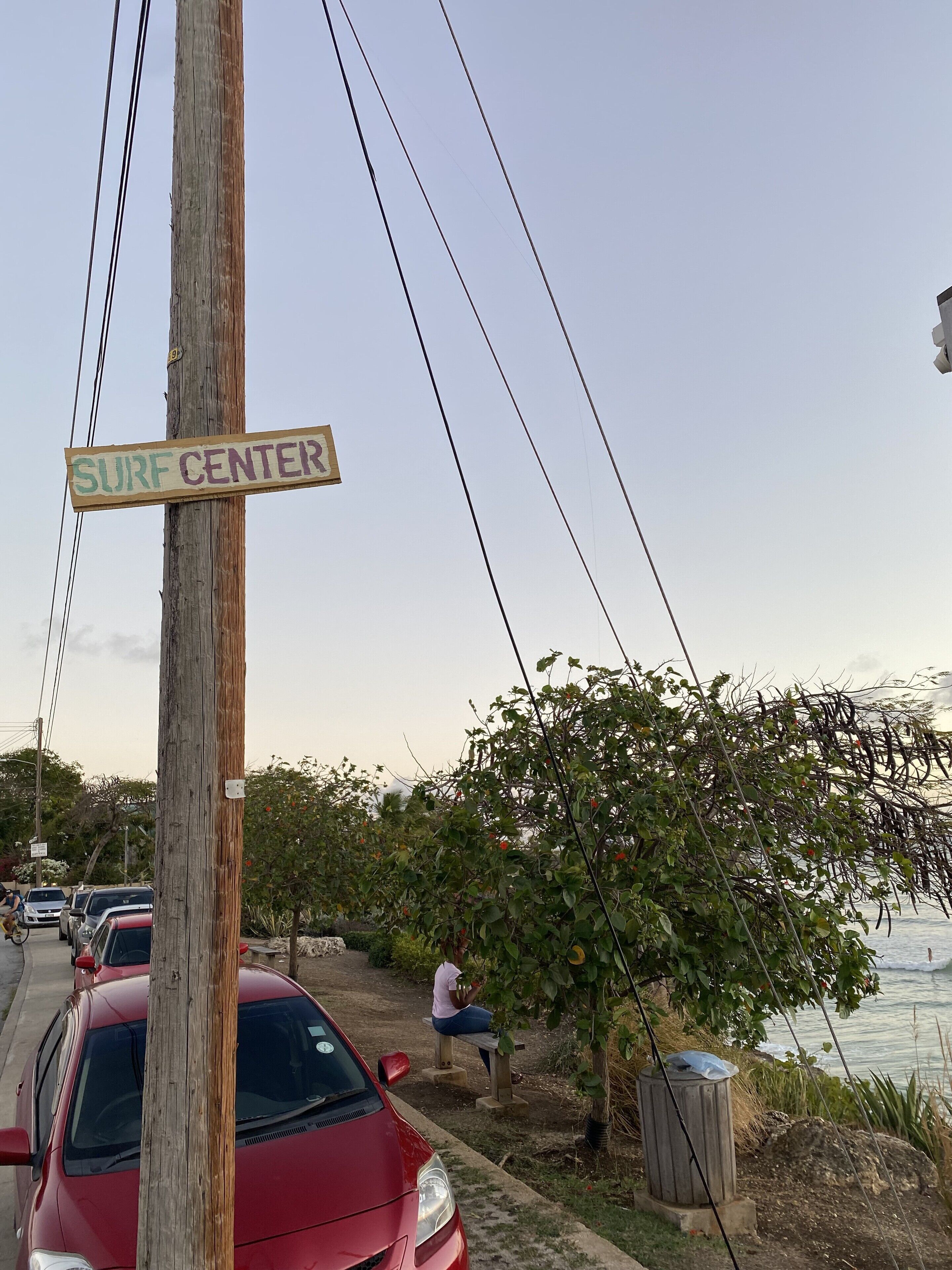 A wooden signpost with "SURF CENTER" written on it, a red car parked beside it, and a person sitting on a bench near a body of water.