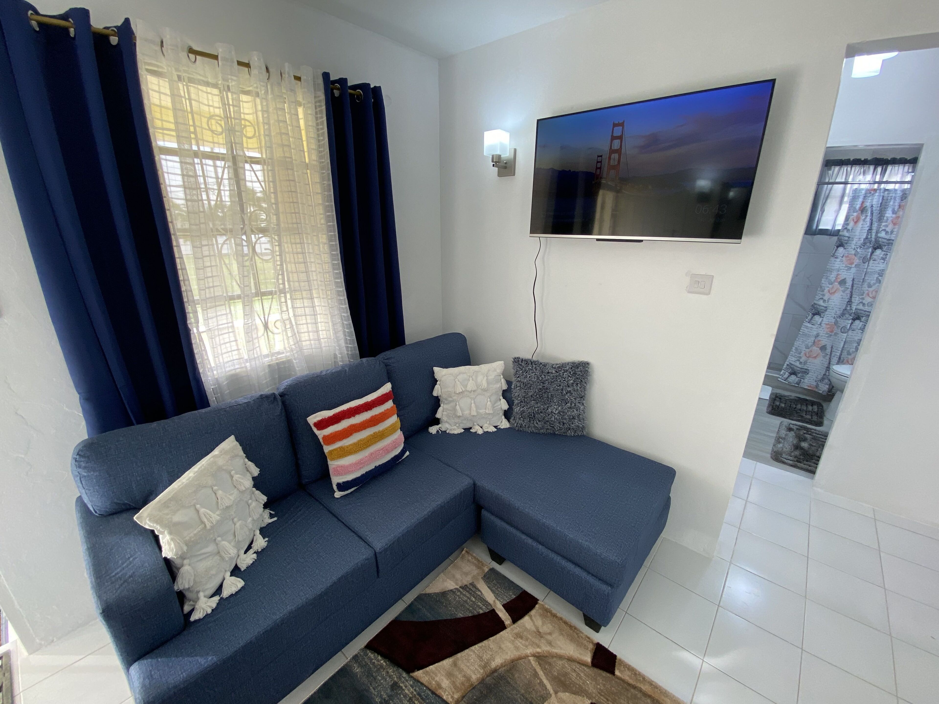 Una sala de estar con un sofá azul, un televisor de pantalla plana montado en la pared y una ventana con cortinas azules.