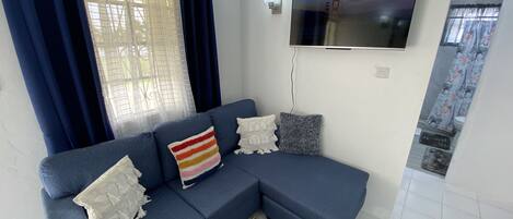Una sala de estar con un sofá azul, un televisor de pantalla plana montado en la pared y una ventana con cortinas azules.