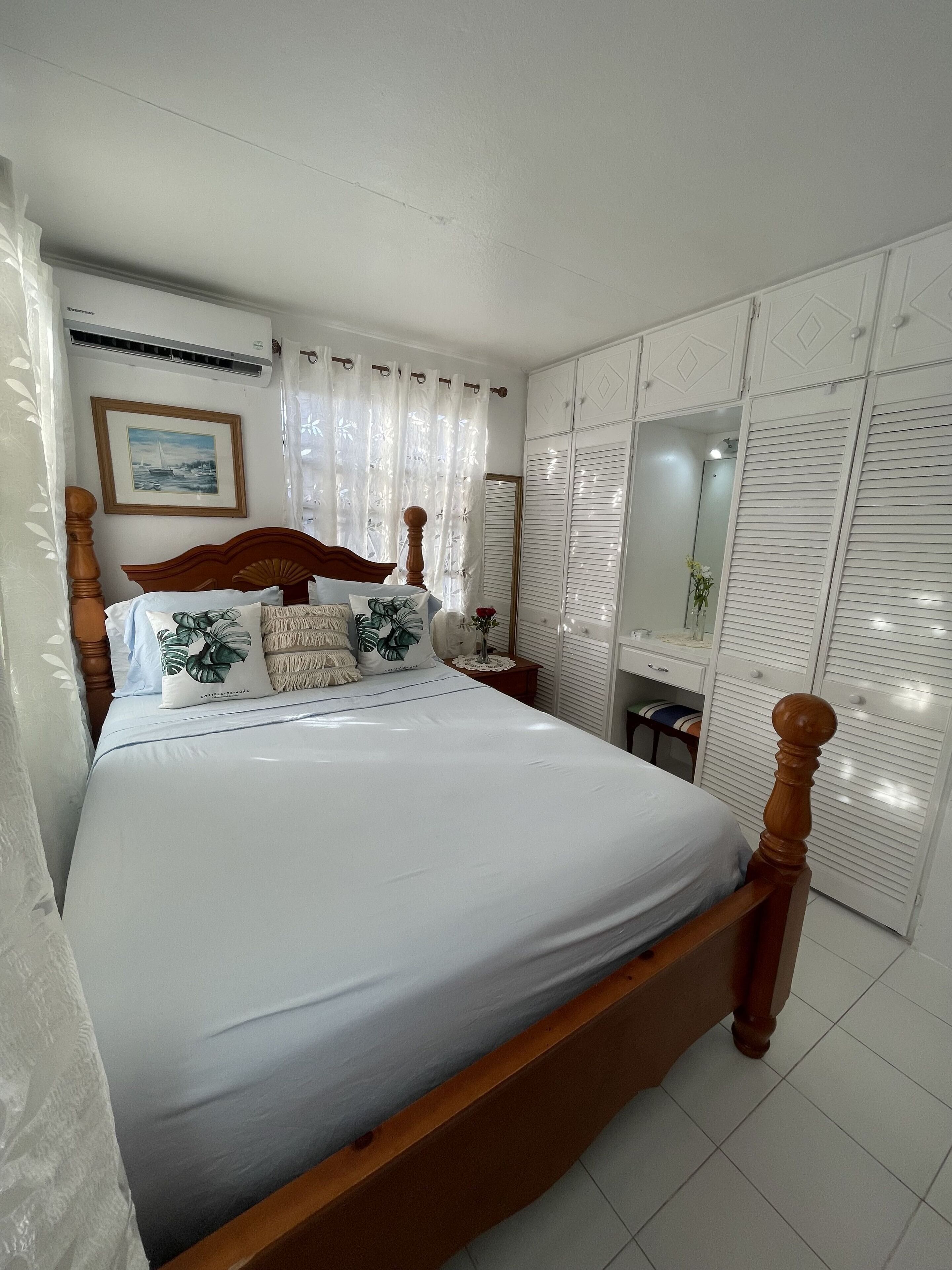 Cama bem arrumada com lençóis brancos, cabeceira de madeira e almofadas decorativas. Há um quadro na parede, uma pequena mesa de cabeceira com um vaso, e um guarda-roupa com portas venezianas.