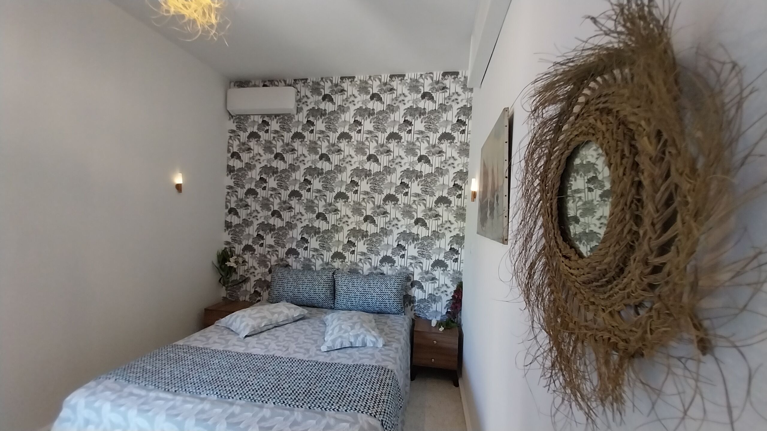 A bedroom with a bed, patterned wallpaper, a wall-mounted air conditioner, and a decorative woven wall piece.