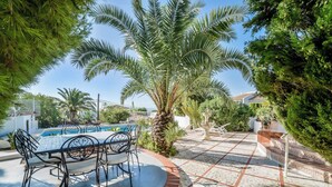 A poolside area with a table and chairs, surrounded by palm trees and a tiled patio.