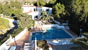 A large, white villa with a swimming pool, surrounded by lush greenery and mountains in the background.
