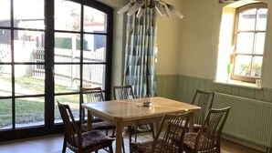 Ein Essbereich mit Holztisch und -stühlen, einem gemusterten Teppich und Blick auf einen eingezäunten Garten durch große Fenster.