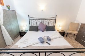 Ein ordentlich bezogenes Bett mit einer weiß-grauen Decke, zwei Handtüchern und einer Fernbedienung. Zwei Nachttische mit Lampen, ein Stuhl und ein Holzschrank sind ebenfalls vorhanden.