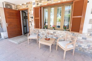 A building entrance with a wooden door, a stone wall, a window with shutters, and two chairs with a table.
