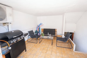 A patio with a grill, two chairs, a table with a plant and drinks, and a TV.