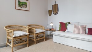 Una habitación con dos sillones de mimbre, un sofá blanco, una mesita auxiliar con una lámpara y un cuadro enmarcado en la pared.