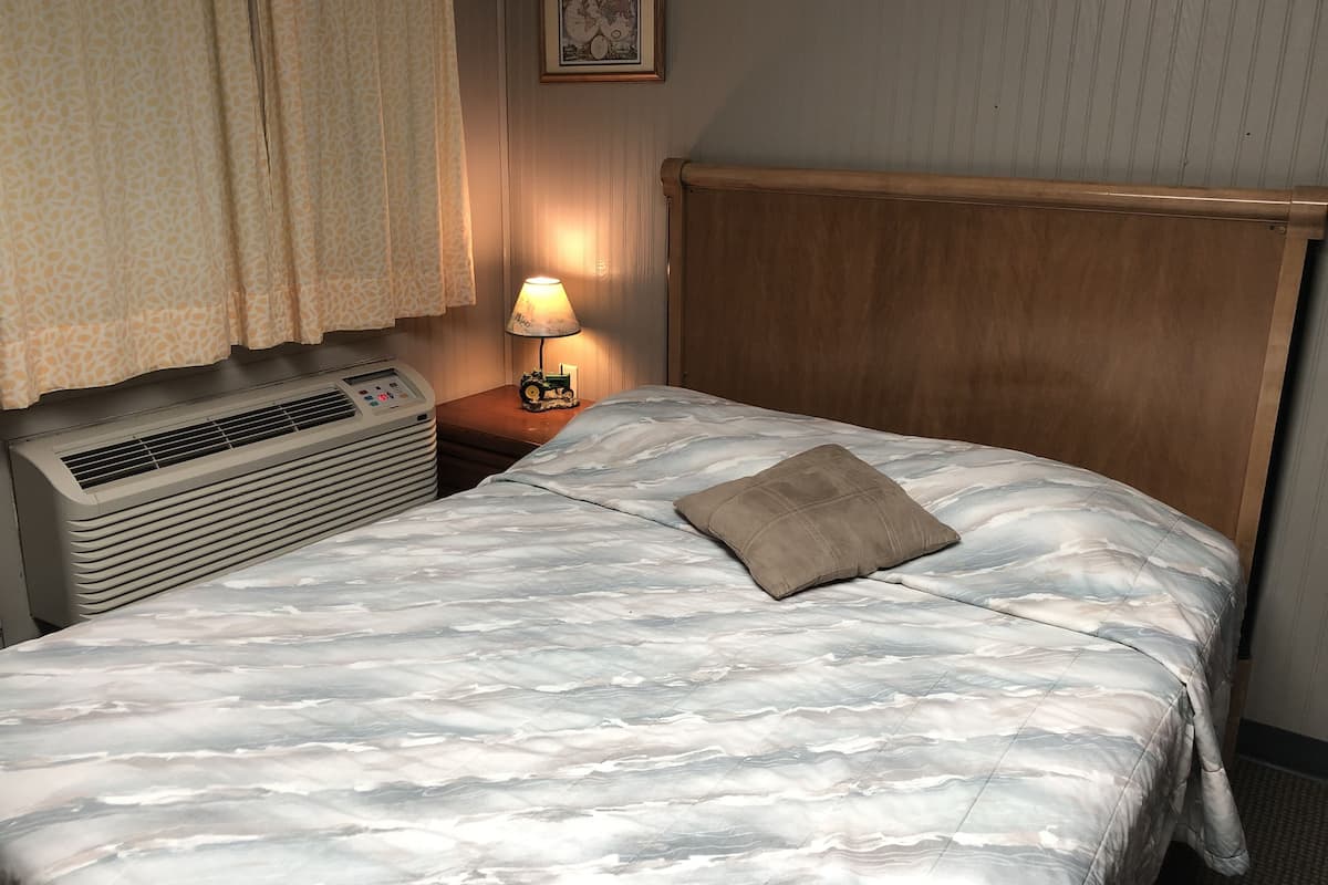 Ein Hotelzimmer mit Bett, Nachttisch mit Lampe, Klimaanlage und einem Fenster mit Vorhängen.