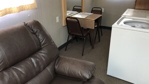 A room with a brown leather sofa, a small table with chairs, a washing machine, and a window with curtains.