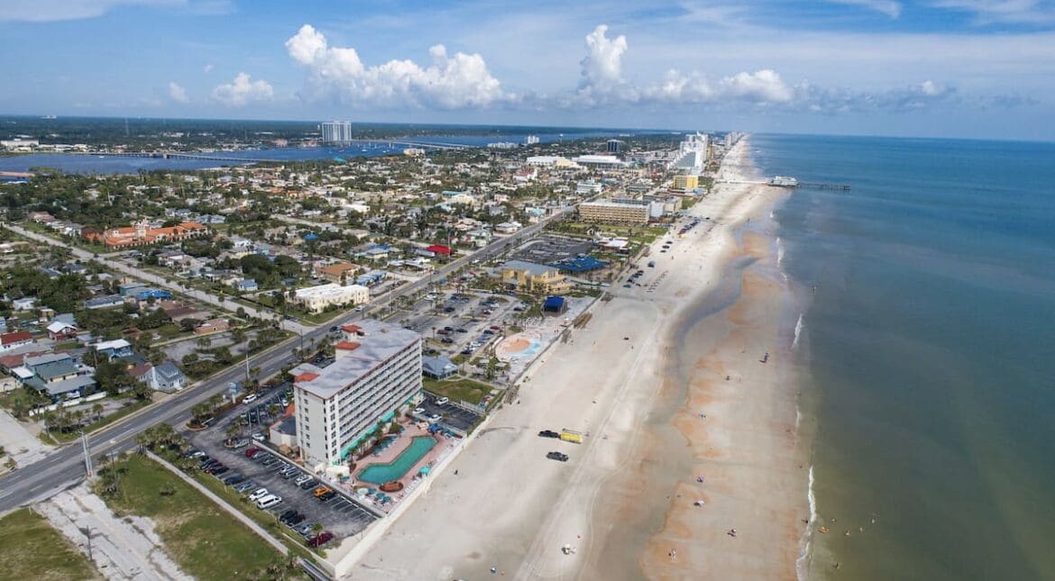 An aerial view of a coastal city with a long beach, a marina, and a large hotel with multiple floors and a swimming pool.