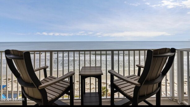 Un balcon avec deux fauteuils Adirondack et une petite table, donnant sur une plage sous un ciel dégagé.