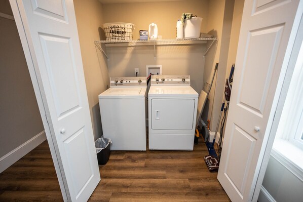 A laundry room with two white washers and dryers, a shelf with laundry supplies, and a broom. - Warm Spring Farmhouse, Old Charm with Modern Updates in Historic Chambersburg (Chambersburg)
