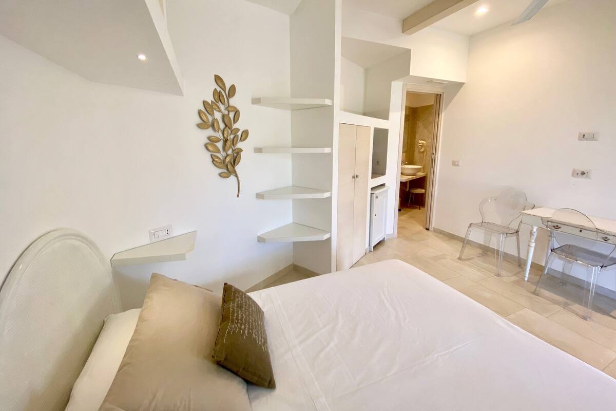 A bedroom with a bed, a desk, and a chair. There is a bathroom visible in the background.