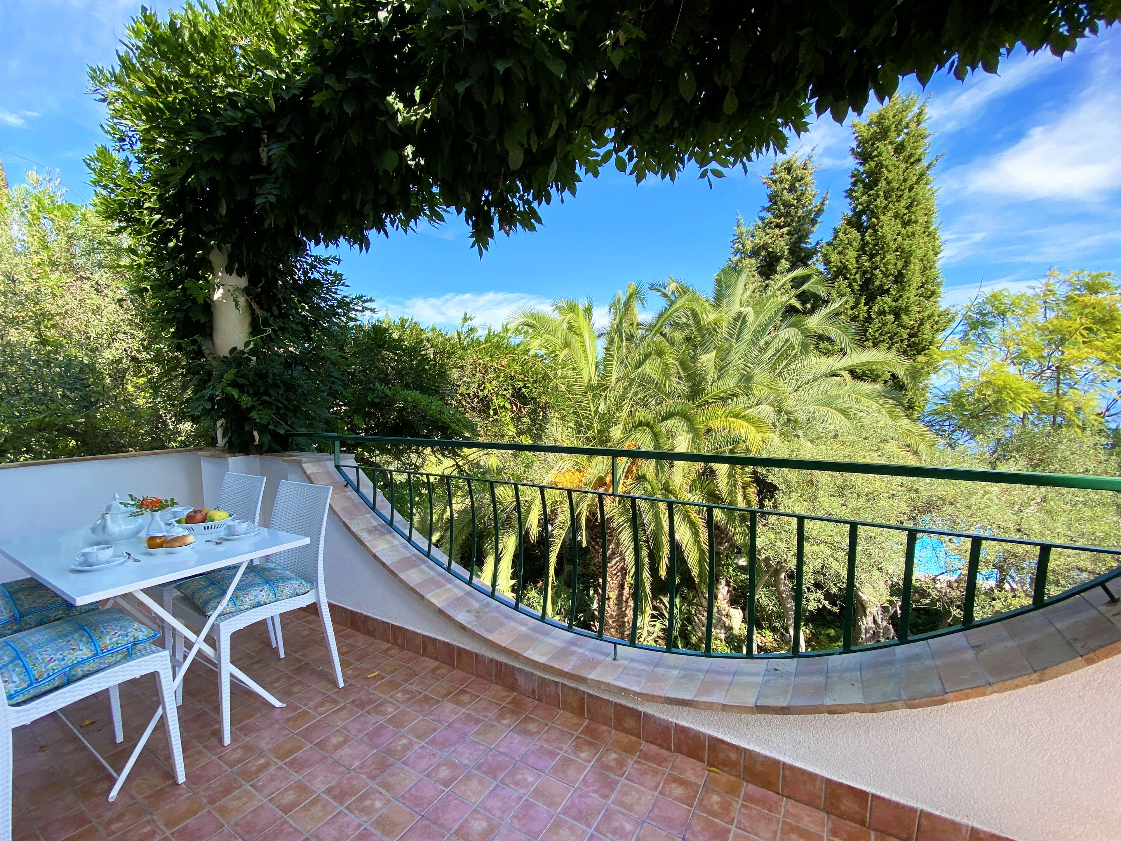 Una terraza con mesa y sillas blancas, rodeada por una barandilla verde, con vistas a un jardín exuberante con varios árboles y plantas.
