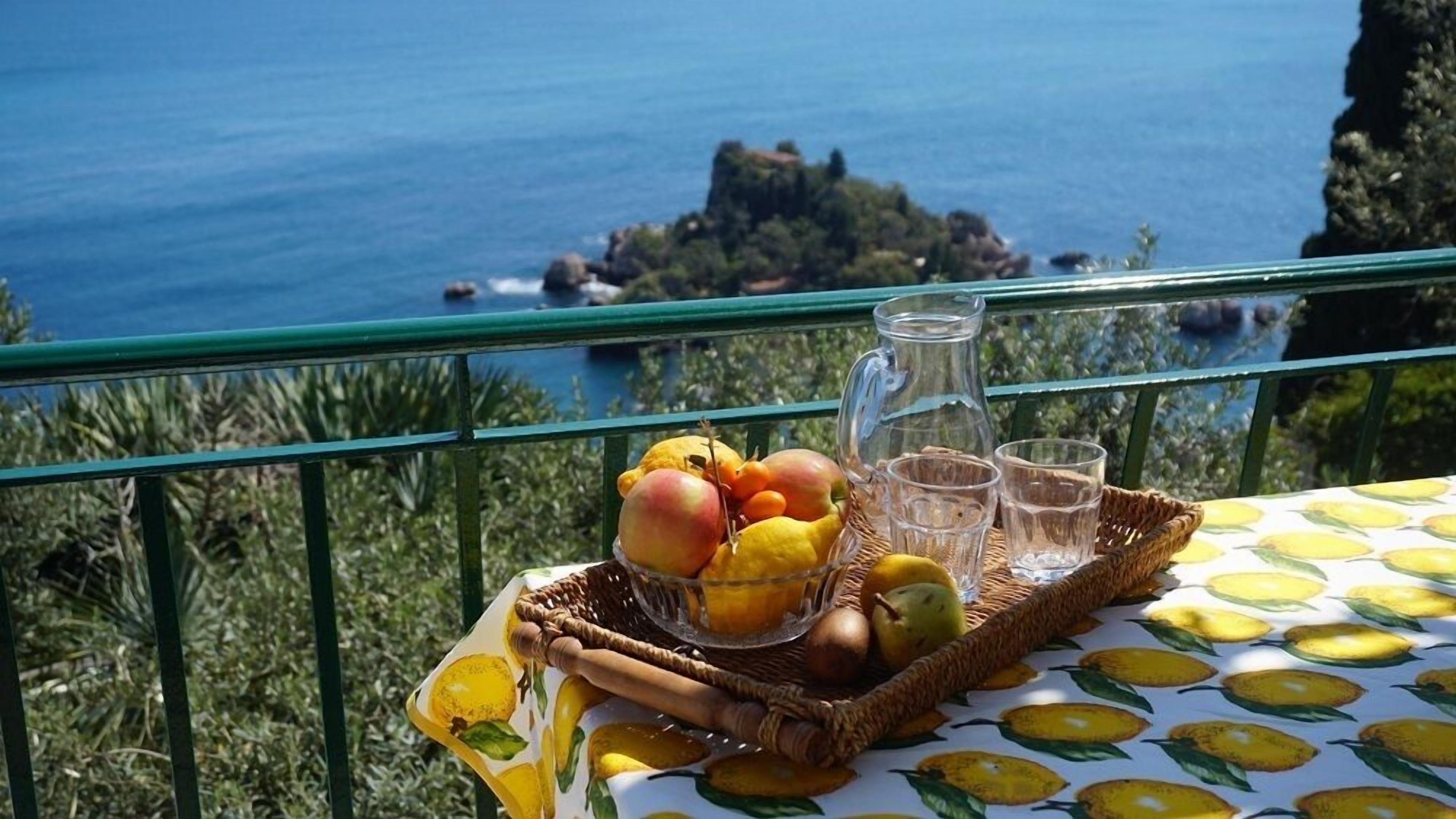 Une table avec un panier de fruits, une carafe et des verres, le tout placé devant un paysage côtier pittoresque.