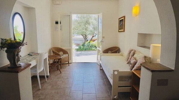 A compact room with a bed, a chair, a small table, and a doorway leading to an outdoor area with a tree.