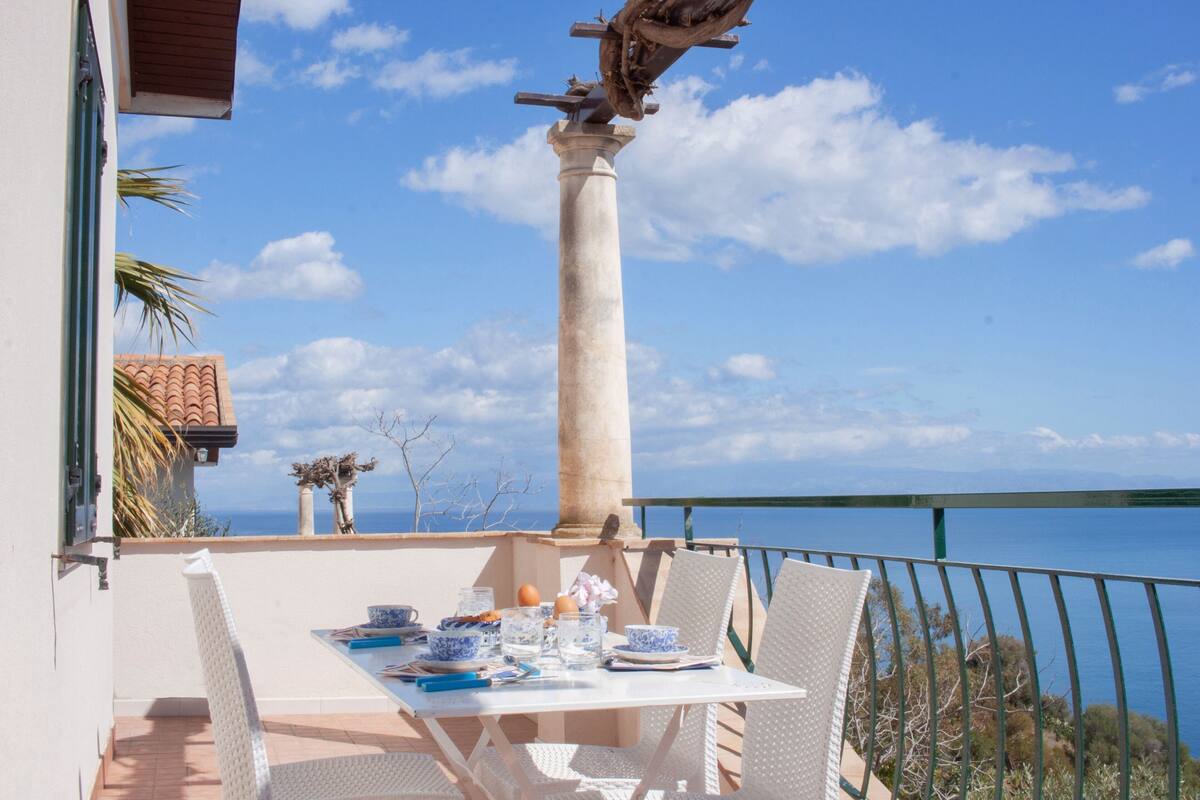 A dining table set with plates, bowls, and glasses on a balcony overlooking the sea.