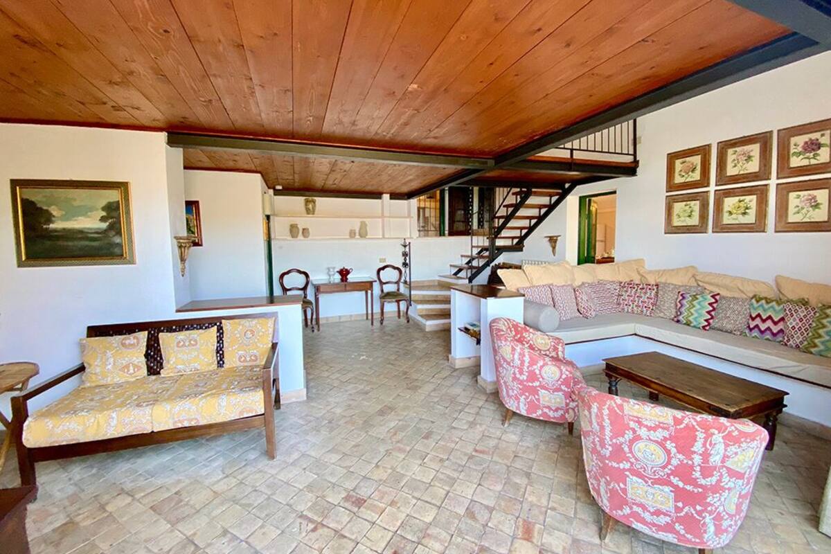 A spacious living area with a wooden ceiling, a sofa set, a dining table, and a staircase leading to an upper level.