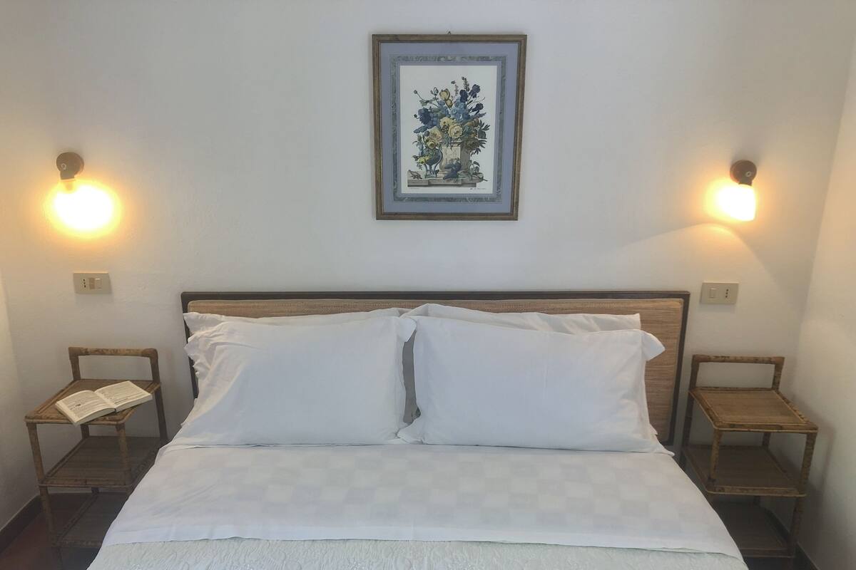A neatly made bed with white linens, two bedside tables with lamps, and a framed botanical print on the wall.