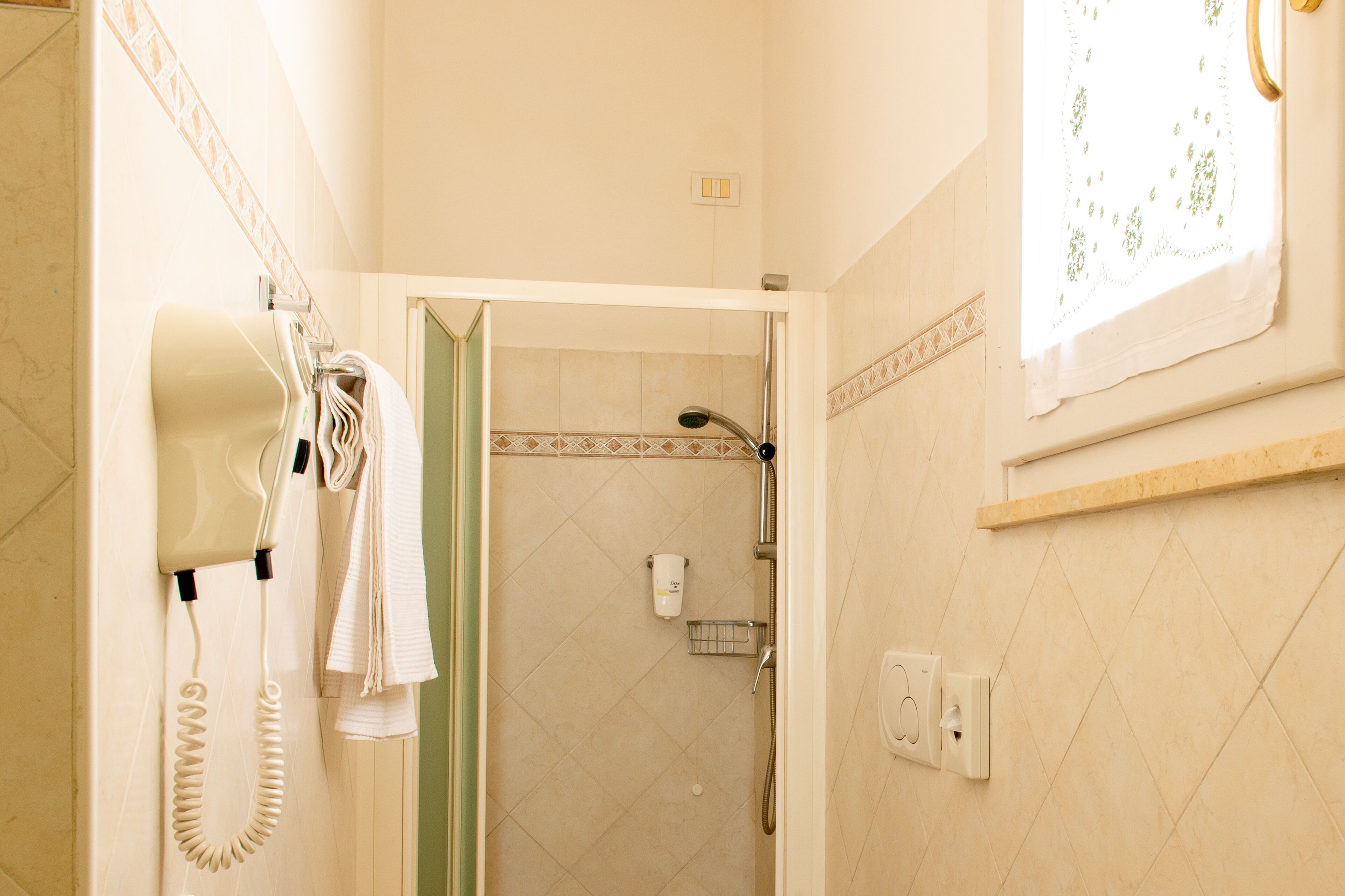 Ein Badezimmer mit Fön, Handtuchhalter, Duschbereich und einem Fenster mit dekorativem Vorhang.