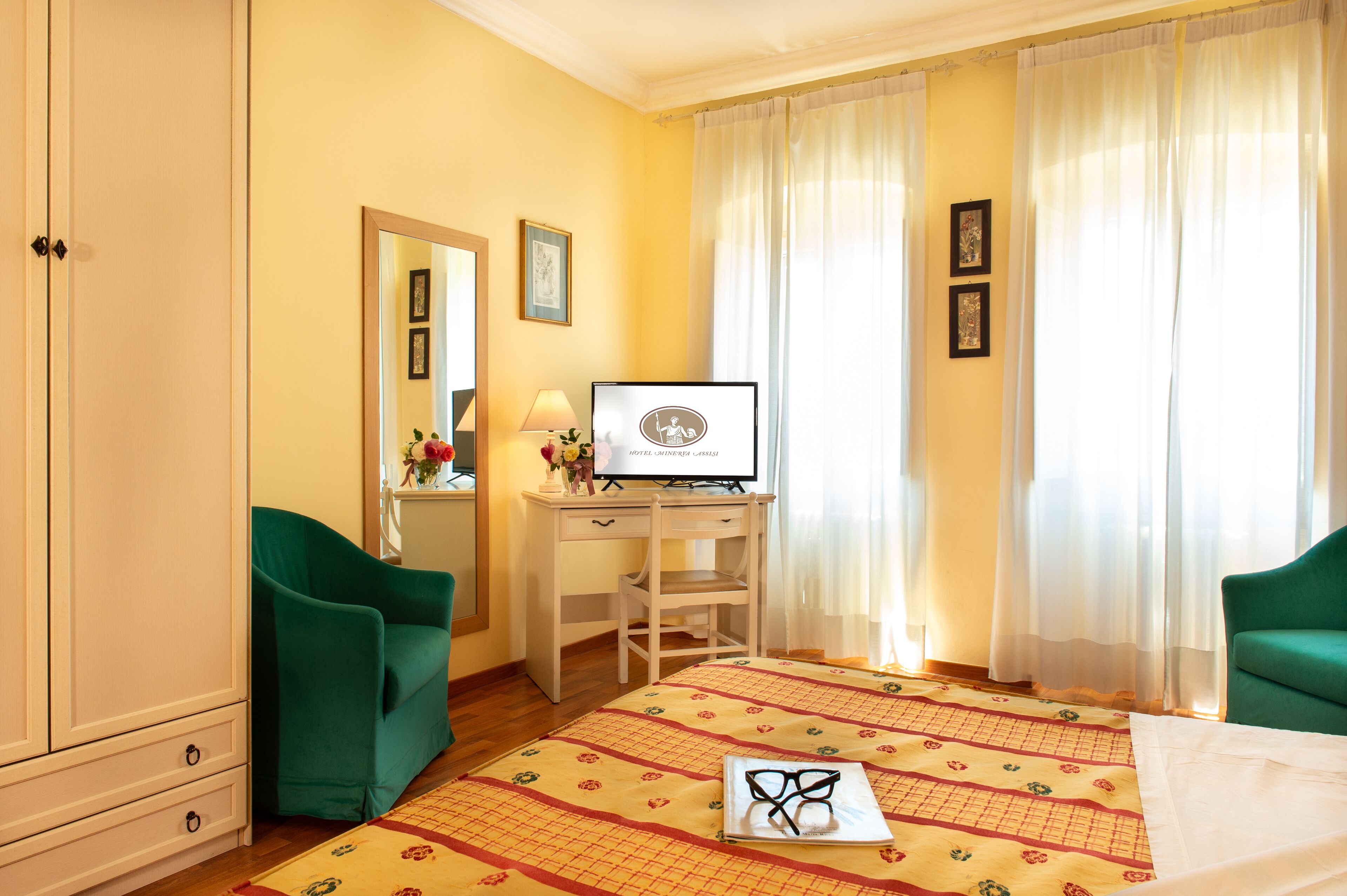 Quarto de hotel com uma cama, uma secretária com computador, duas cadeiras verdes, um guarda-roupa e cortinas.
