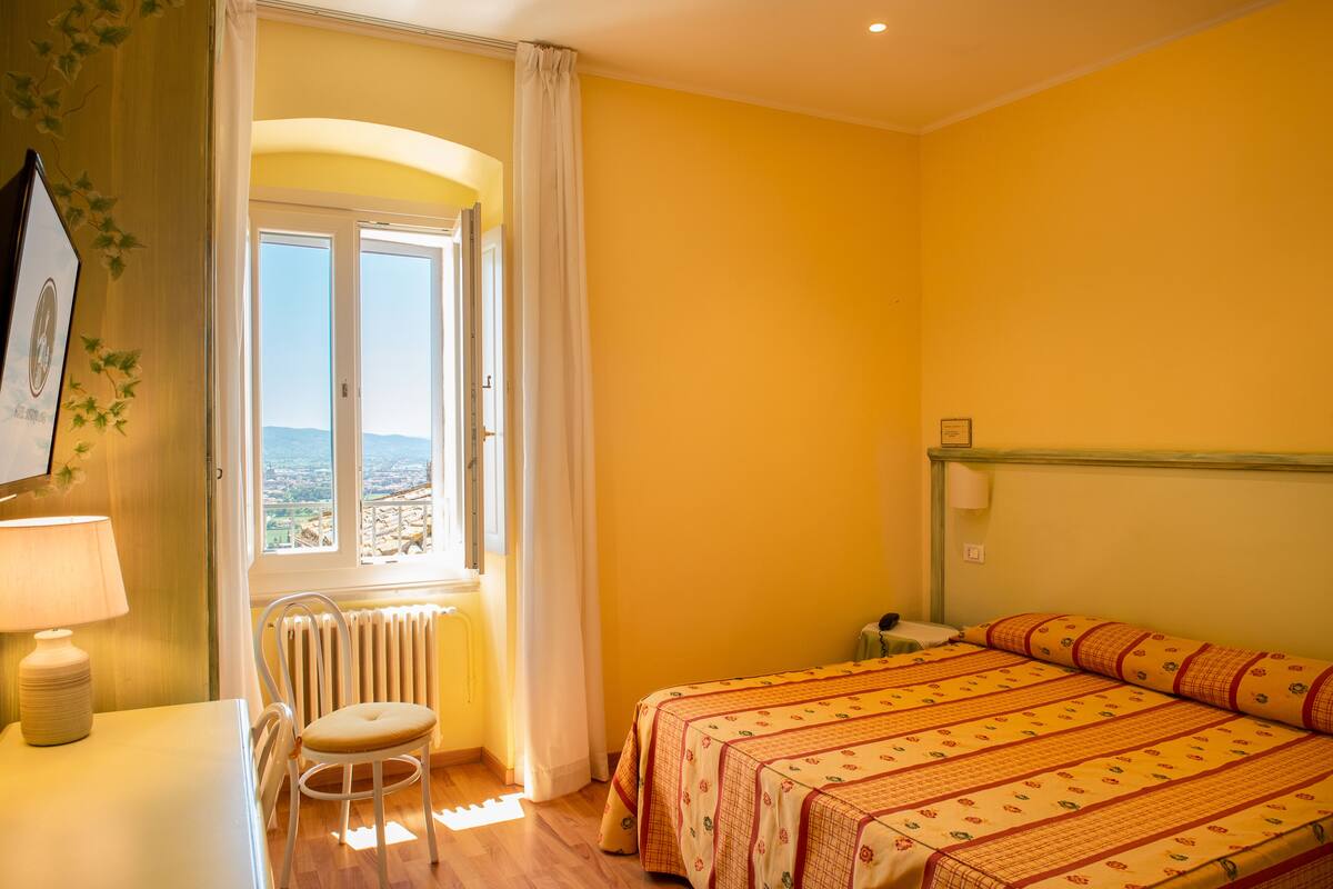 A room with a bed, a chair, a table, and a window with a view.