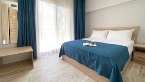 Une chambre bien rangée, avec un lit, des tables de chevet, un banc et une grande fenêtre avec des rideaux.