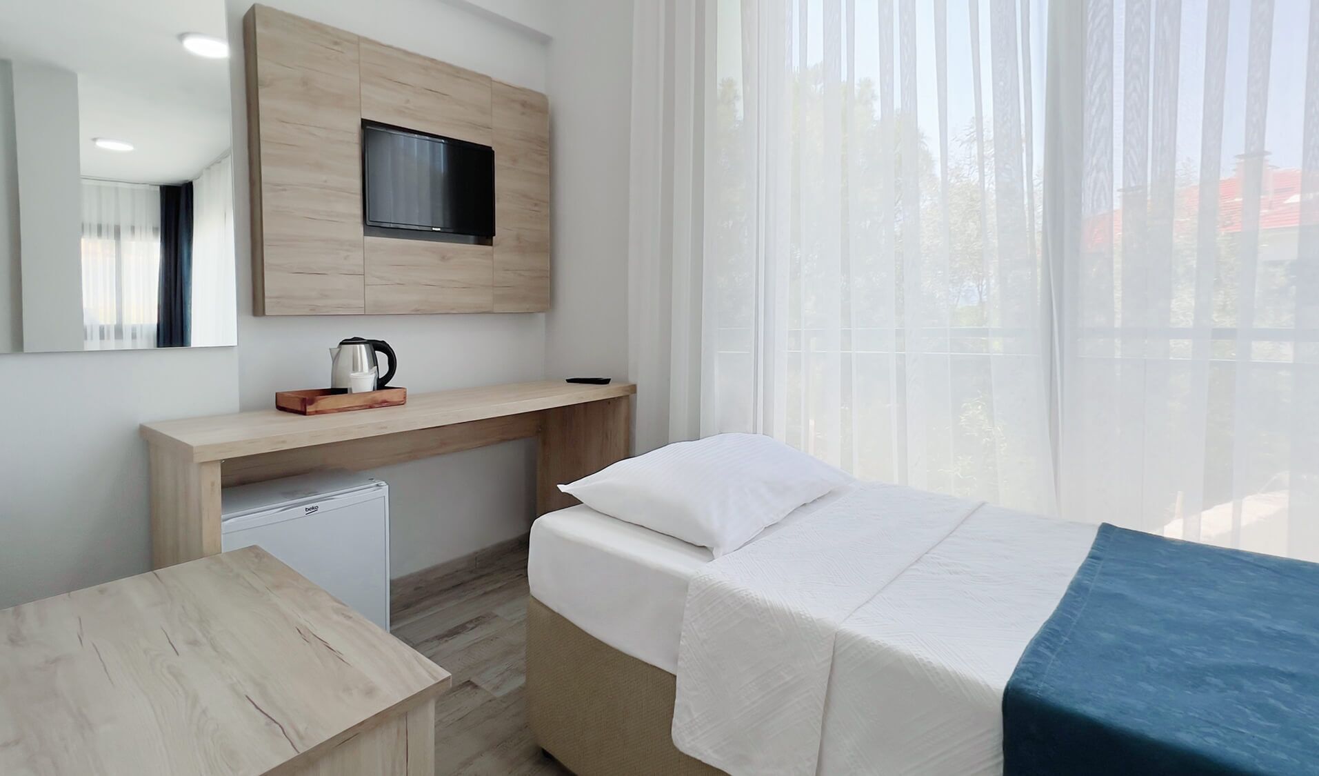 A hotel room with a bed, a desk, a small refrigerator, a television mounted on the wall, and a window with sheer curtains.
