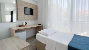 A hotel room with a bed, a desk, a small refrigerator, a television mounted on the wall, and a window with sheer curtains.