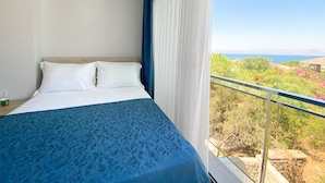 Une chambre à coucher avec un lit, une fenêtre avec des rideaux et un balcon offrant une vue sur la verdure et la mer.