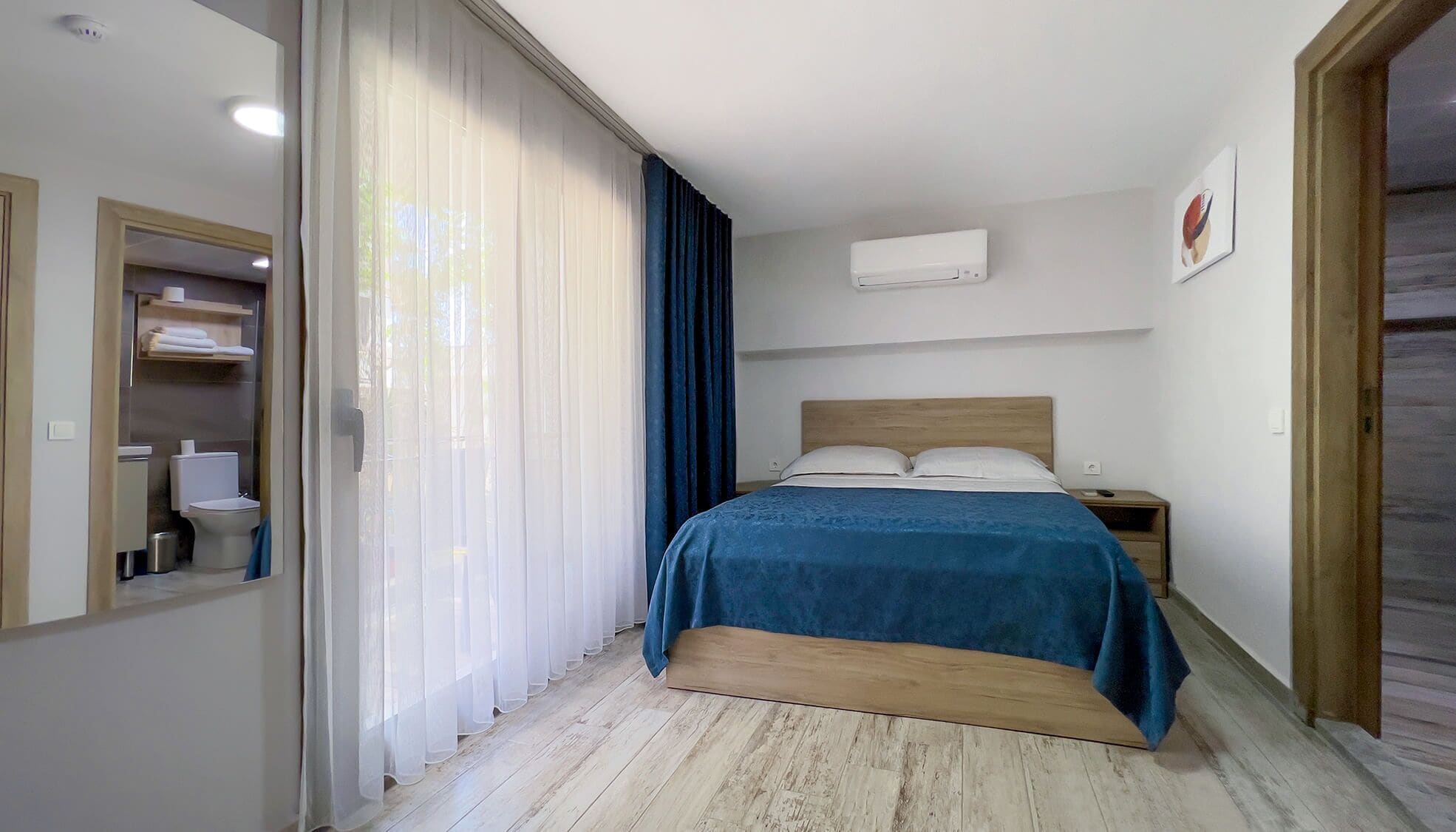 A bedroom with a bed, a nightstand, a wall-mounted air conditioner, and a bathroom with a toilet and sink.