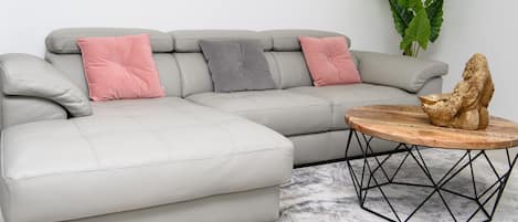 A light grey sectional sofa with three cushions in pink, grey, and pink. A round wooden coffee table with a gold sculpture sits in front.