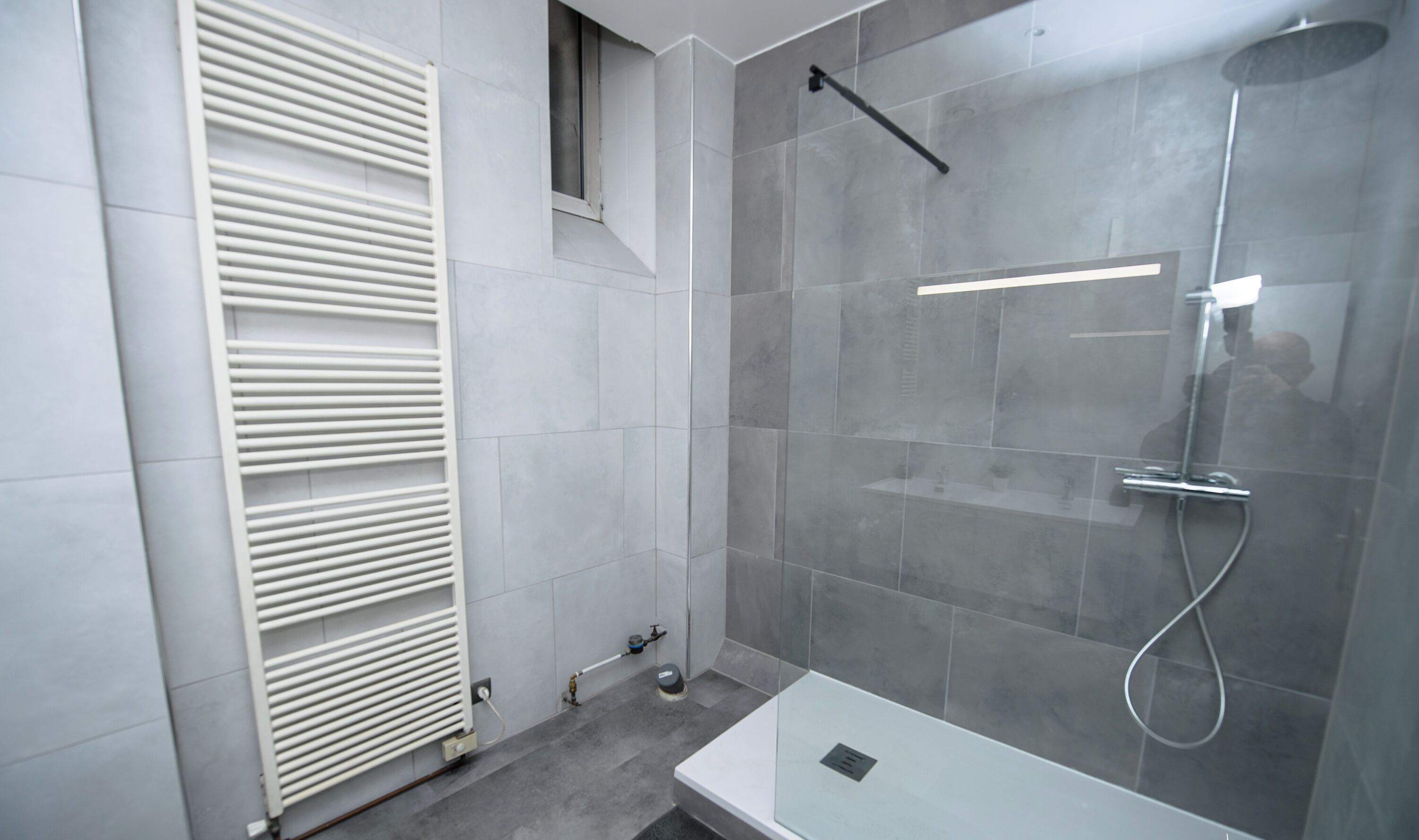A modern bathroom with a glass shower enclosure, a white radiator, and a small window.